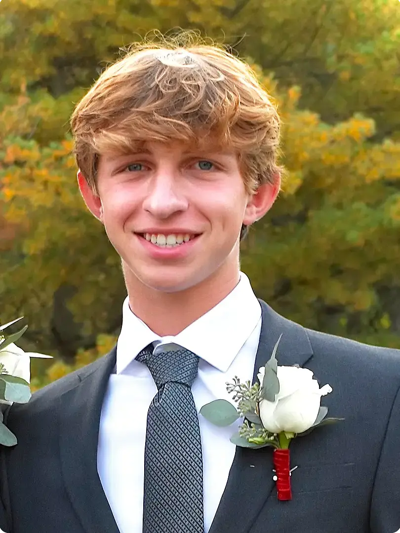Mitchell in a suit and tie smiles outdoors, wearing a white boutonniere. Autumn foliage is visible in the background, creating a warm, cheerful atmosphere.
